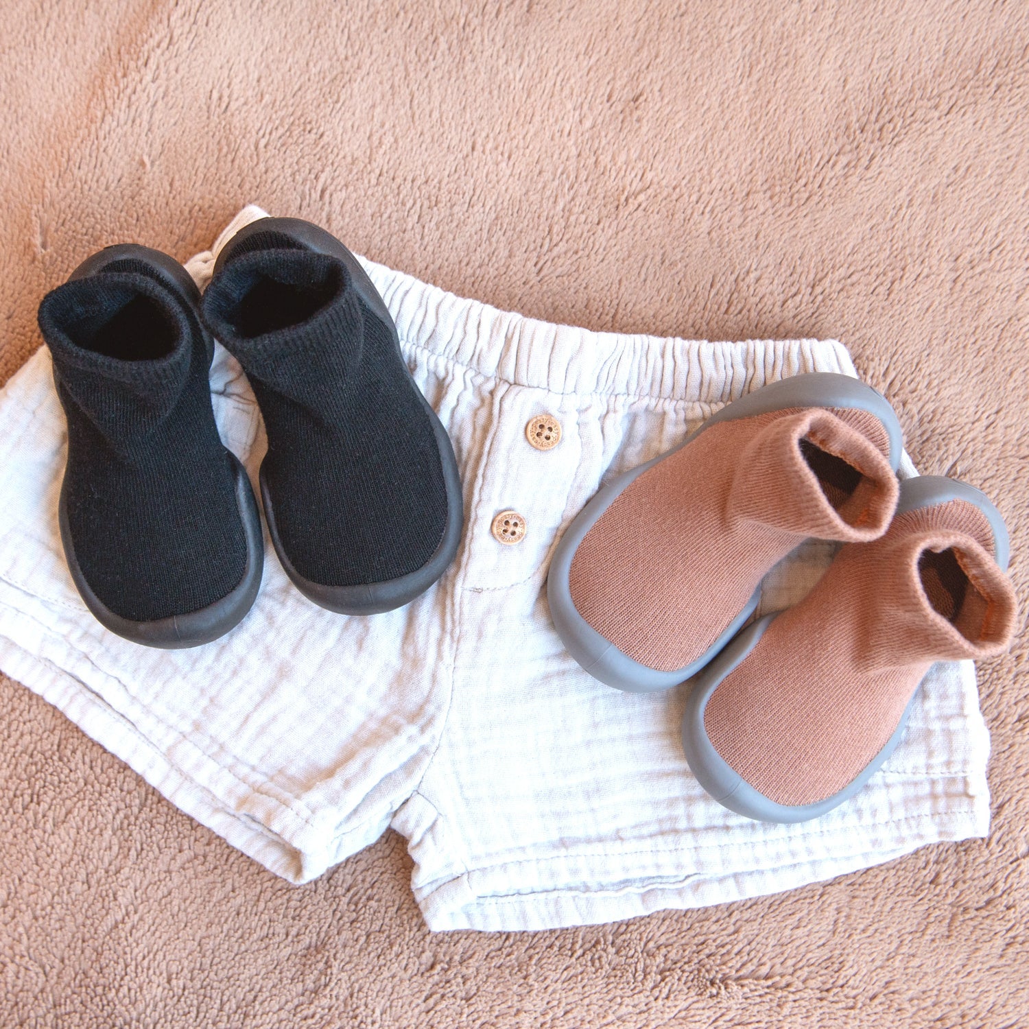 A pair of tan baby shoes, a pair of black baby shoes, and white linen shorts on a fuzzy tan blanket.