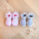 Two pairs of baby shoes, pink and gray, with white lace trim, on a wooden floor with baby's breath.