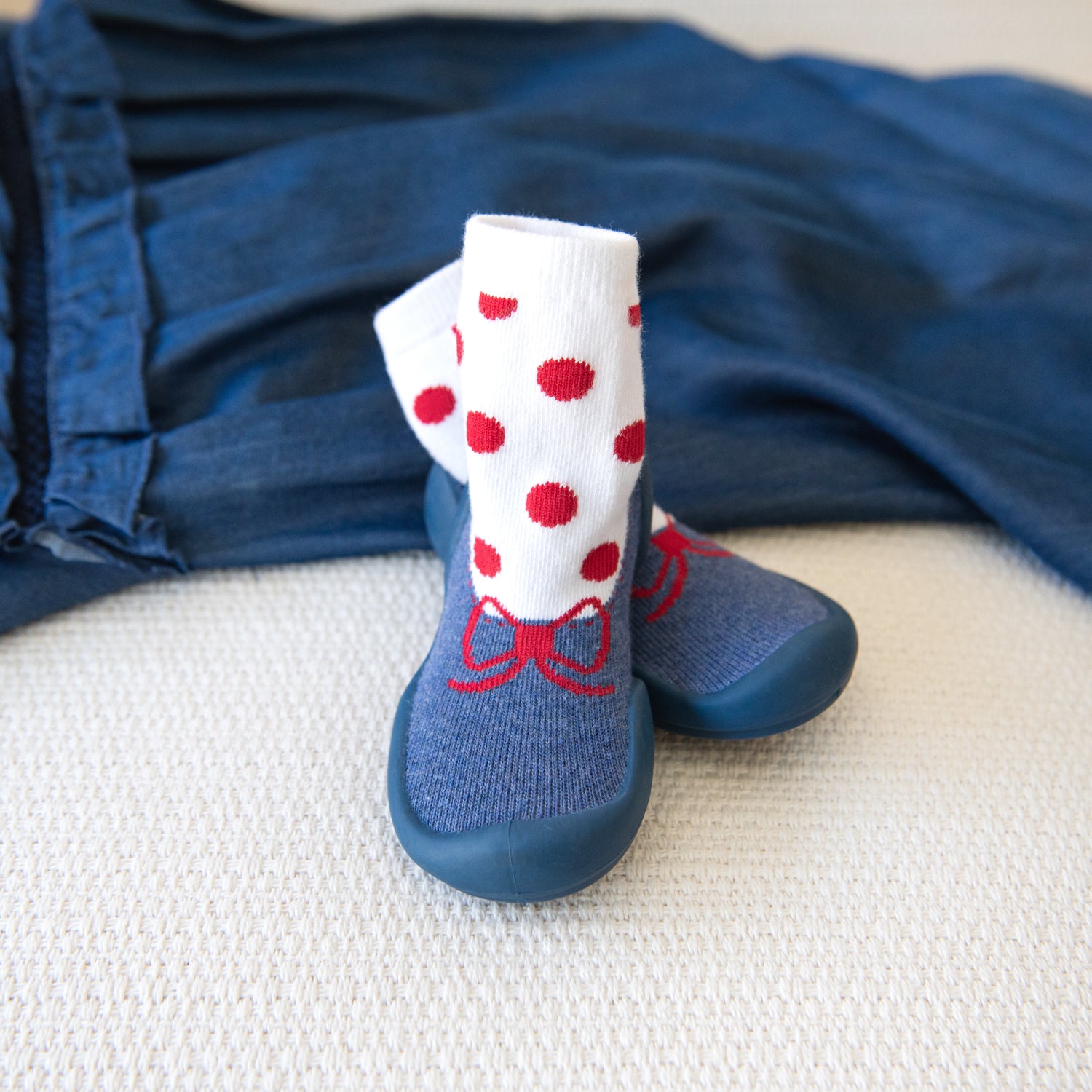 Two baby sock shoes, blue bottoms, blue socks with red bow, and white socks with red dots, on a white textured surface with a blue shirt in the background.