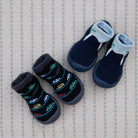 Two pairs of baby sock-shoes, one black with colorful car pattern, the other dark blue with light blue accents, on a textured light beige surface with a faint vertical stripe.