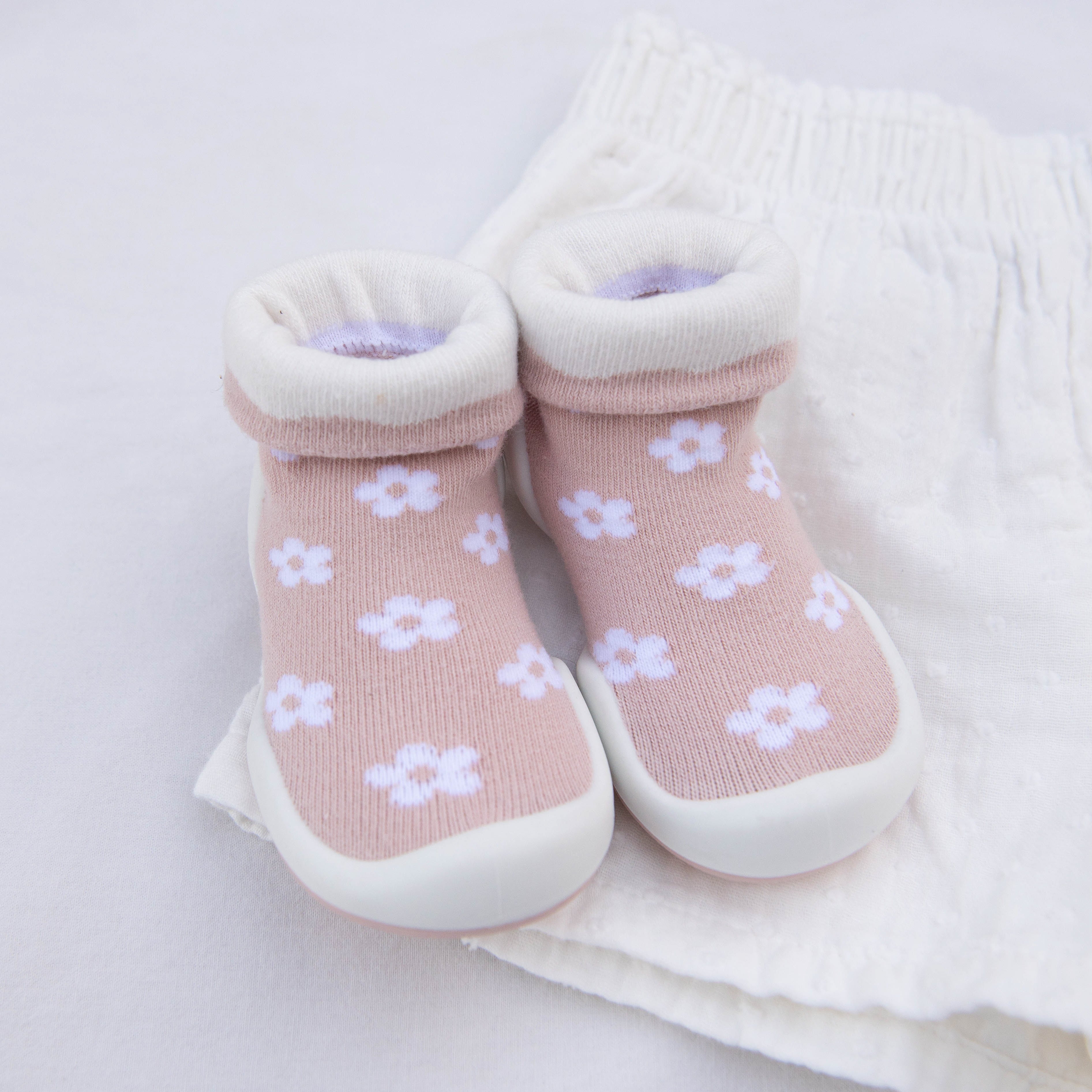 Close-up of pink baby socks with white flowers, rolled white cuffs, and white rubber soles, sitting on a white fabric surface.