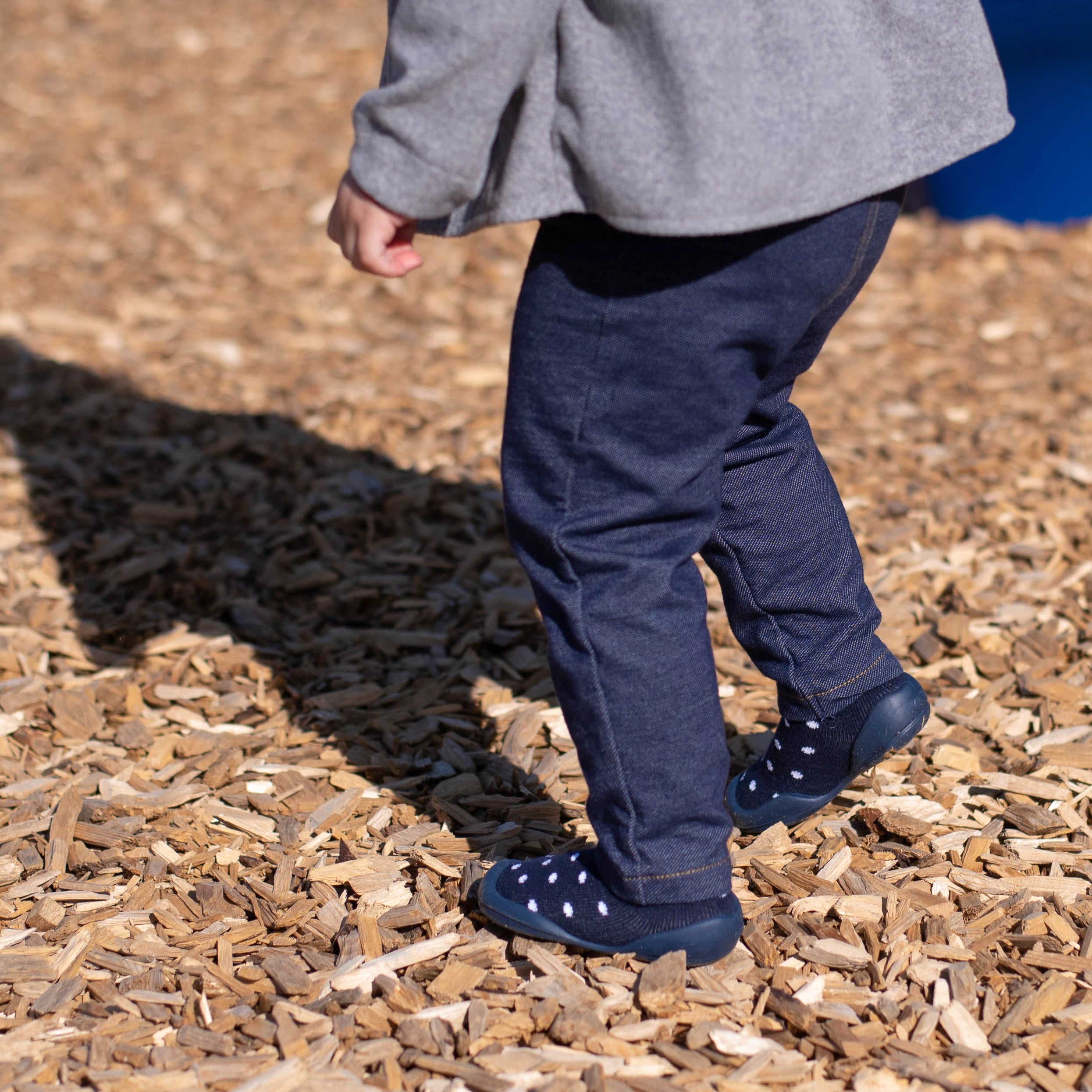 Toddler in gray sweater and blue denim pants walks on wood chips in blue and white polka dot sock shoes.