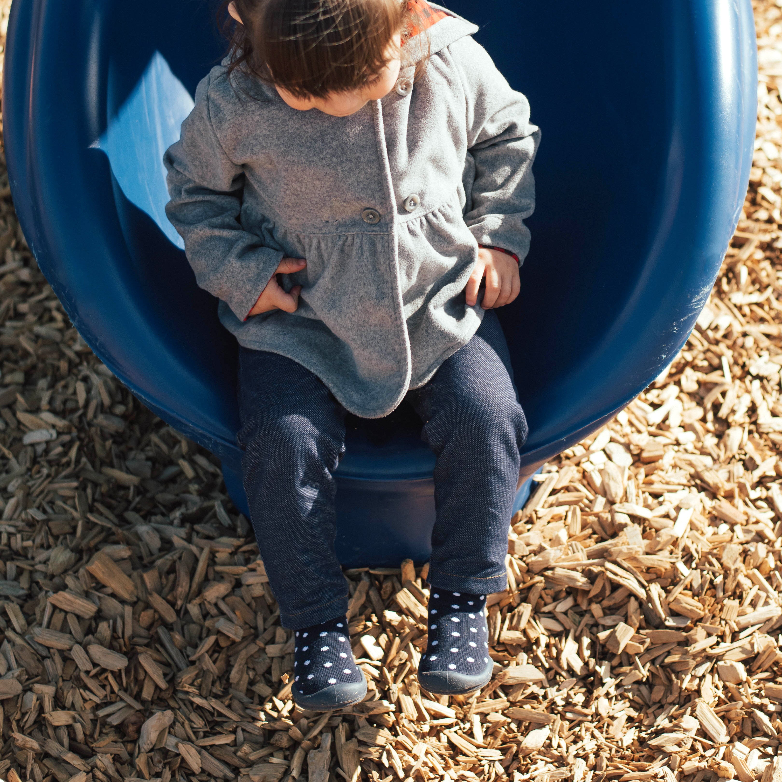 Child sitting on a blue slide, wearing a gray coat, blue pants, and polka dot socks, surrounded by wood chips.