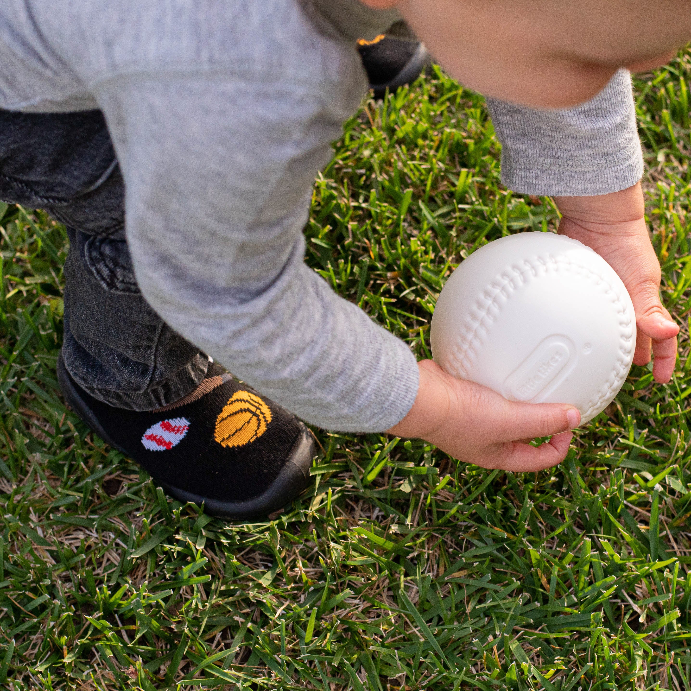 Toddler in grey shirt, dark jeans, and basketball socks picking up a white plastic baseball on green grass.