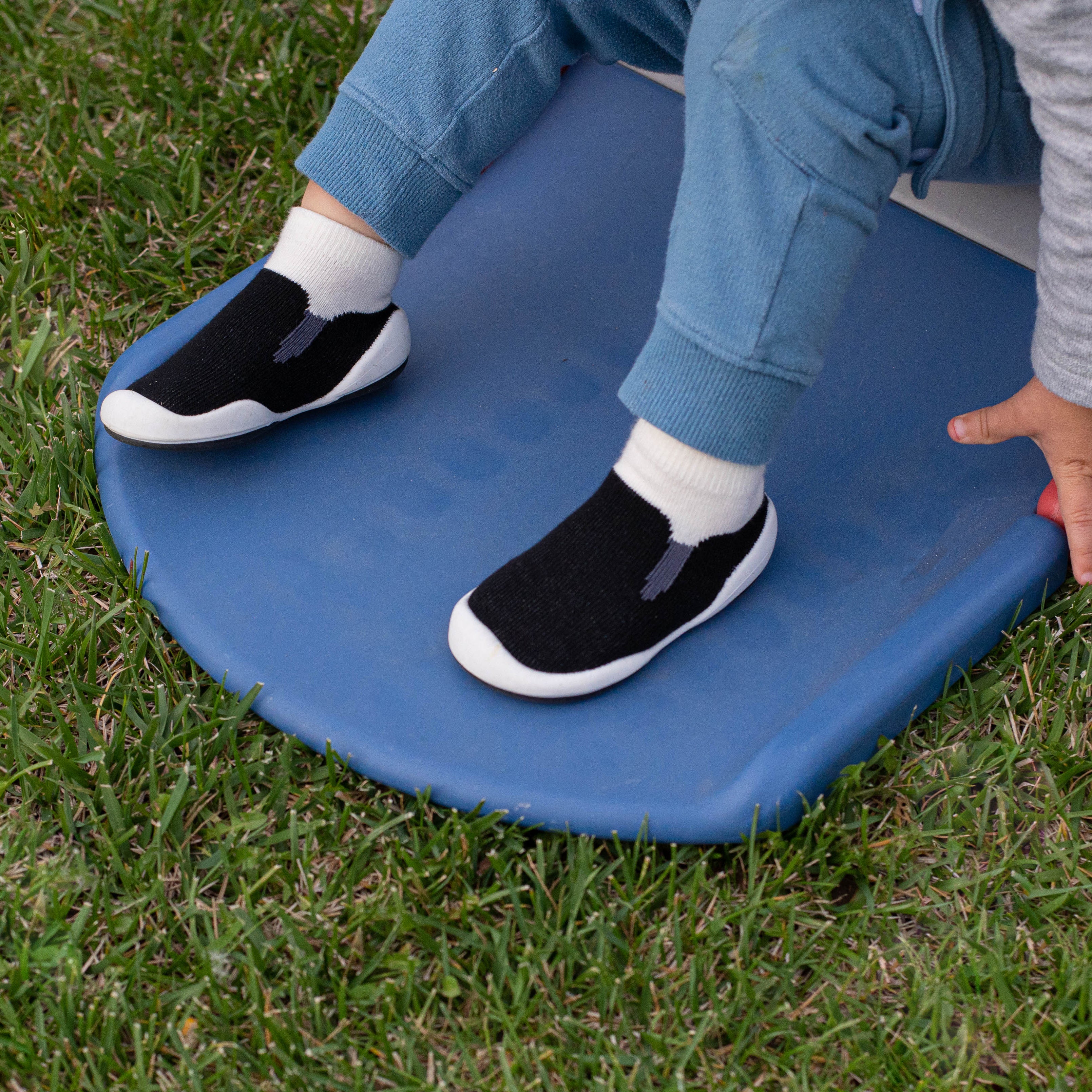 A child with blue pants and black shoes sits on a blue sled on the grass.