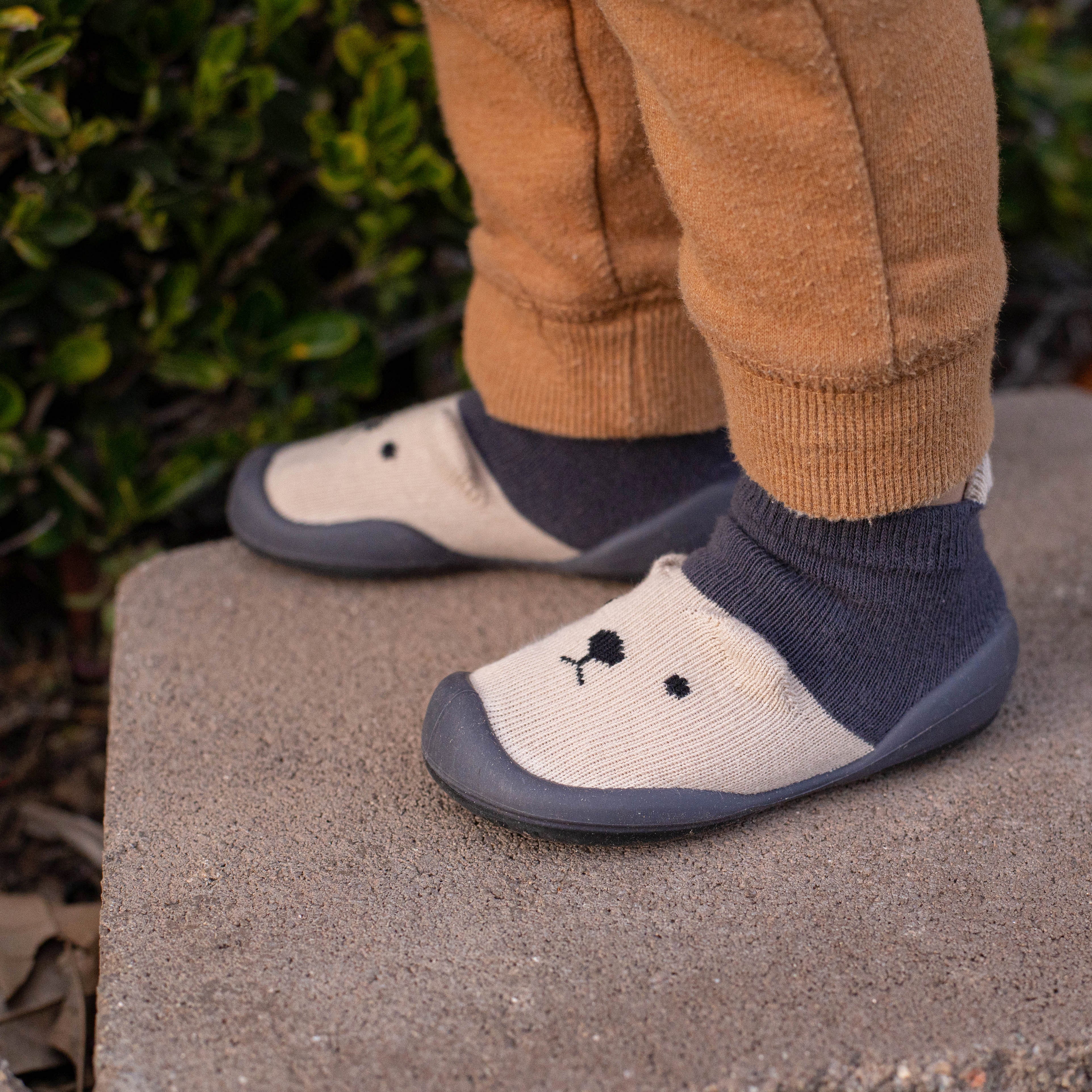 A toddler wearing tan pants and gray-and-beige sock shoes with a black bear design standing on a gray concrete step.