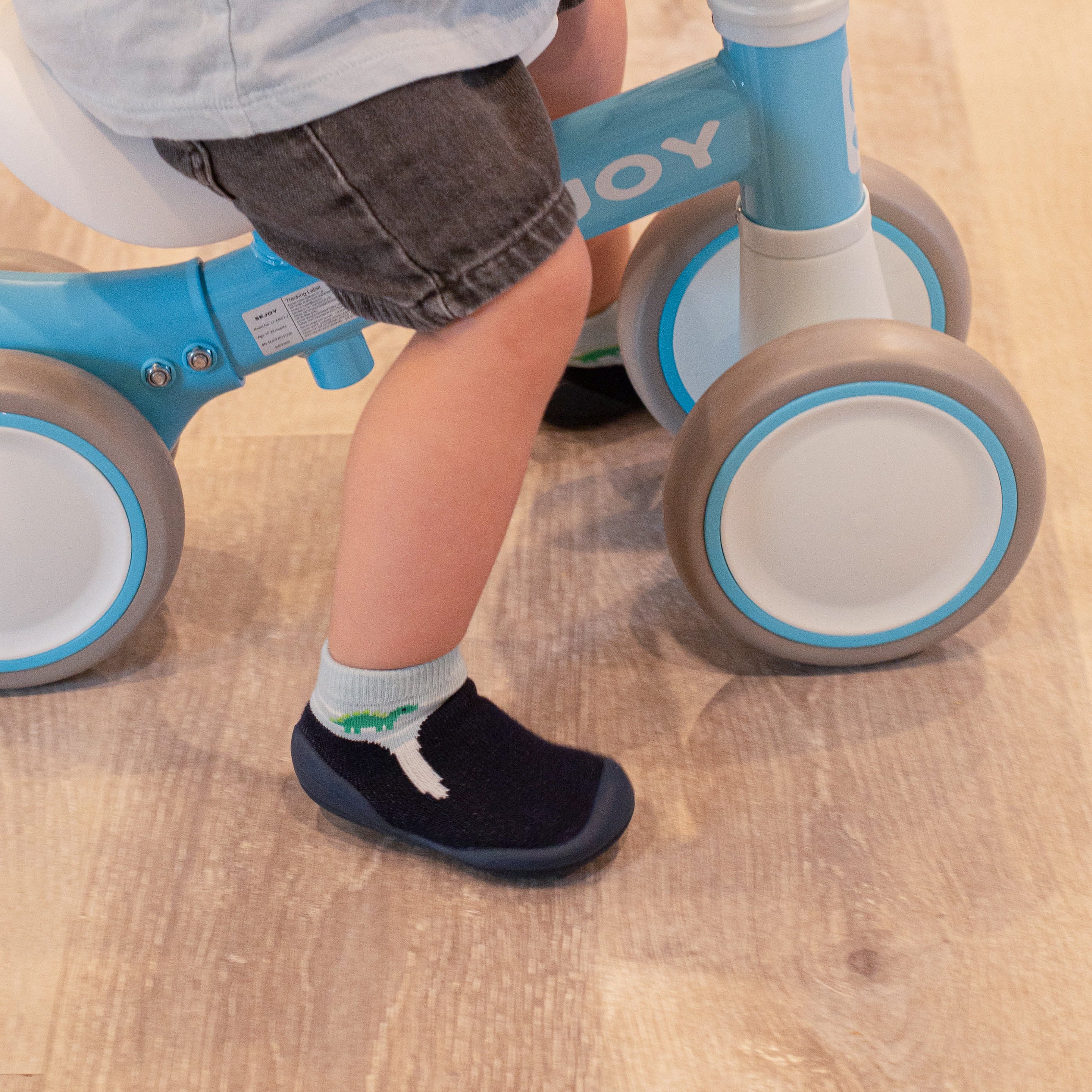 Child riding a blue balance bike while wearing gray shorts, a light blue shirt, and dark blue socks with a dinosaur pattern.