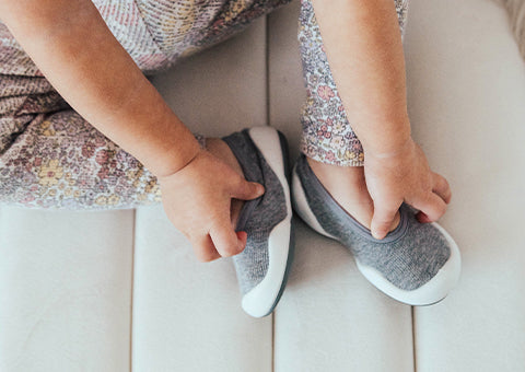 Child wearing Komuello gray flat sock shoes with white soles on a light sofa.