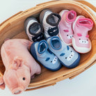 A wooden basket containing a pink pig plush toy and three pairs of baby shoes: blue hippo, pink pig, and gray and white cat.