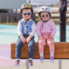 Two toddlers in helmets and sunglasses sit on a wooden bench.
