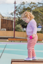 A toddler in pink sweater and pants standing on a bench, holding a pink toy.