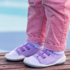 Toddler wearing pink corduroy pants and lavender knit shoes with white socks, standing on wood planks.