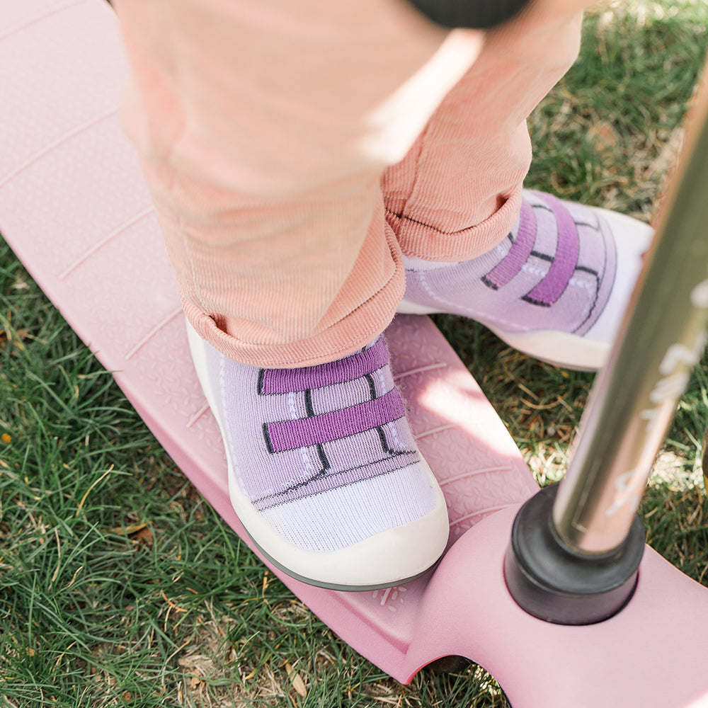 Child in peach pants and lavender shoes standing on a pink balance board in grass.