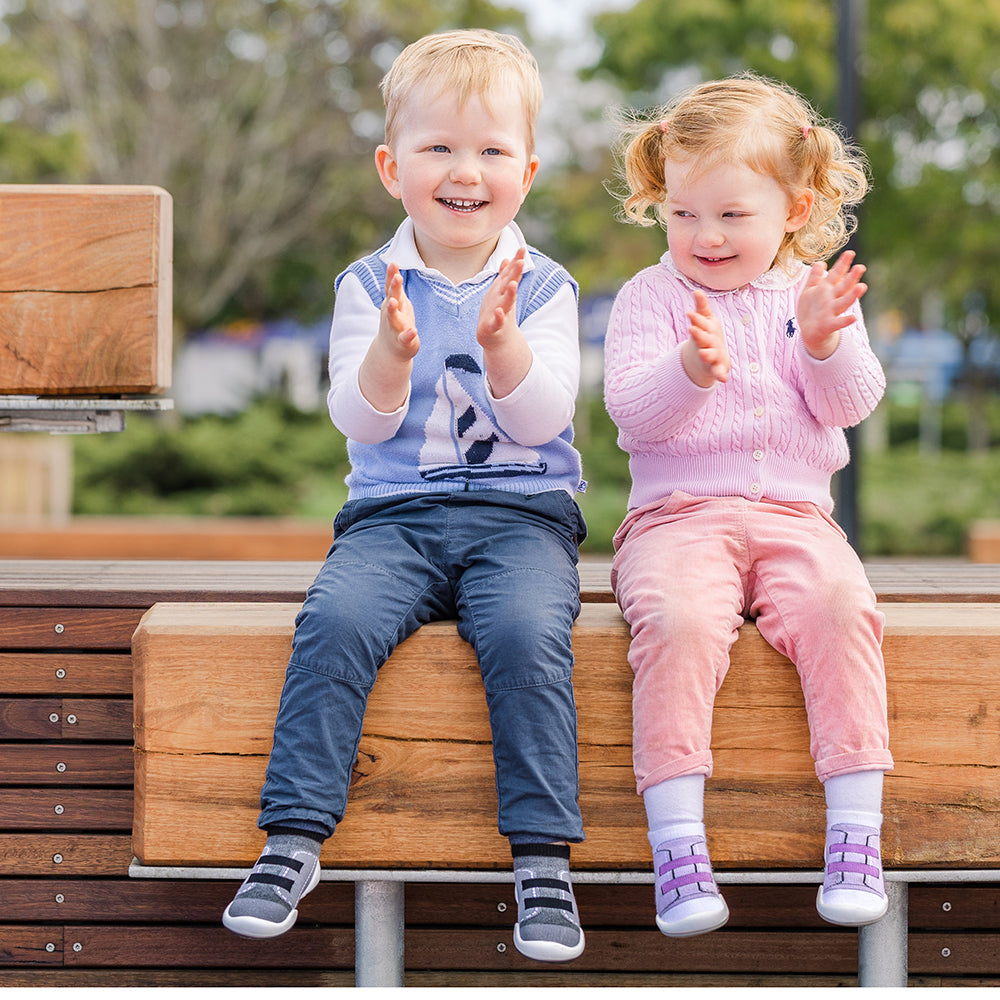 Two young children in casual outfits sitting on a wooden bench and clapping.