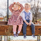 Two children sitting on a wooden bench, girl in a floral dress and leggings, boy in a sweater vest and jeans.