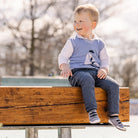 A blond toddler sits on a wooden bench, wearing a blue sweater vest and gray pants.