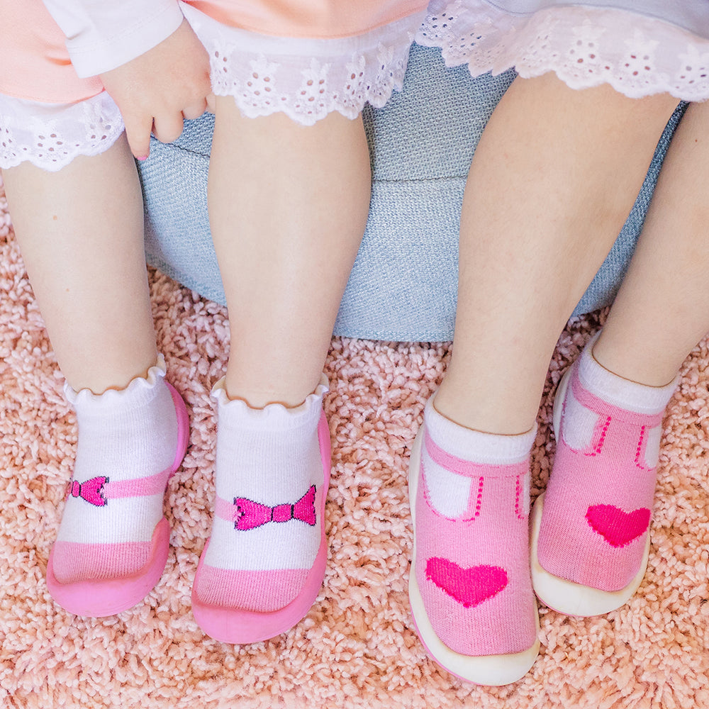 Two children's legs and feet wearing white and pink socks with bow and heart designs, sitting on a pink shag rug.
