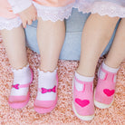 Two children's legs and feet wearing white and pink socks with bow and heart designs, sitting on a pink shag rug.