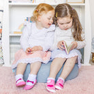 Two young girls in dresses and socks reading a book on a gray cushion in front of a white bookcase on a pink rug.