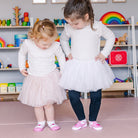 Two young girls in white shirts and tutus, wearing pink shoes.