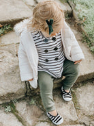 Toddler in striped shirt, green pants, white jacket, and black and white shoes, seated on stone steps.