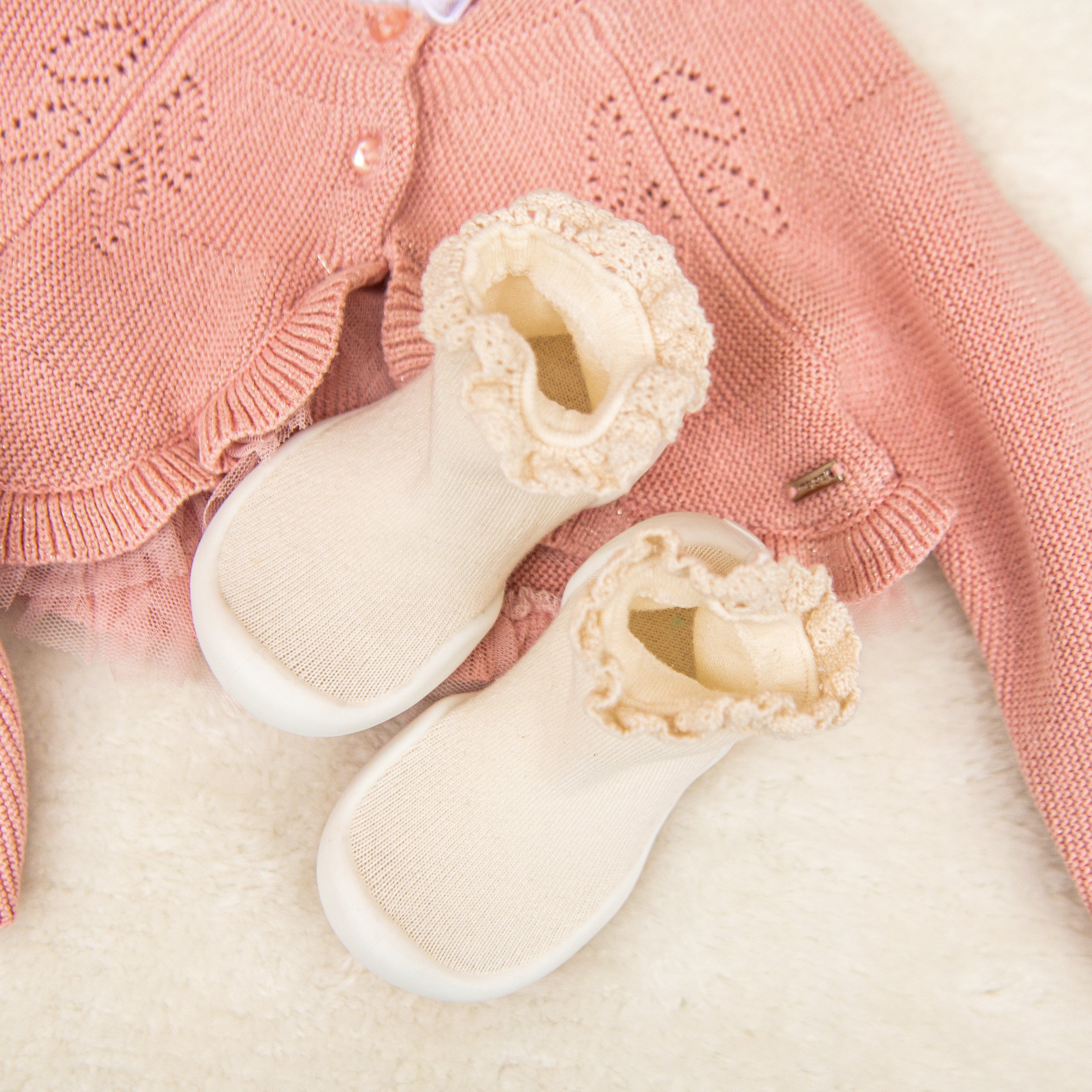 A pink knit cardigan with a pair of white baby shoes on a fluffy white background.