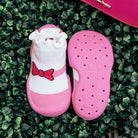 Pink and white baby shoes with a red bow, set on artificial greenery.