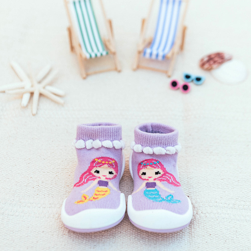 Close-up of two lilac baby socks/shoes with white soles, decorated with mermaid graphics, beach chairs, a starfish, sunglasses, and seashells in the background, set on a white surface.