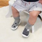 Toddler wearing gray shorts with surfboard pattern, gray socks with star and stripe patterns, and white sole shoes sitting on white stool and fluffy rug.