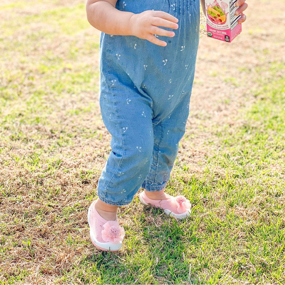 Toddler in blue jumpsuit and pink floral shoes holding juice box on grass.