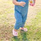 Toddler in blue jumpsuit and pink floral shoes holding juice box on grass.