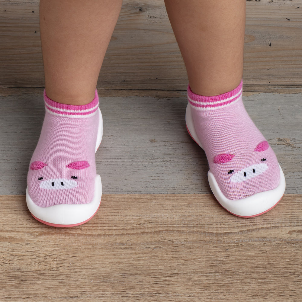 Child's pink pig-themed sock shoes on wooden floor.
