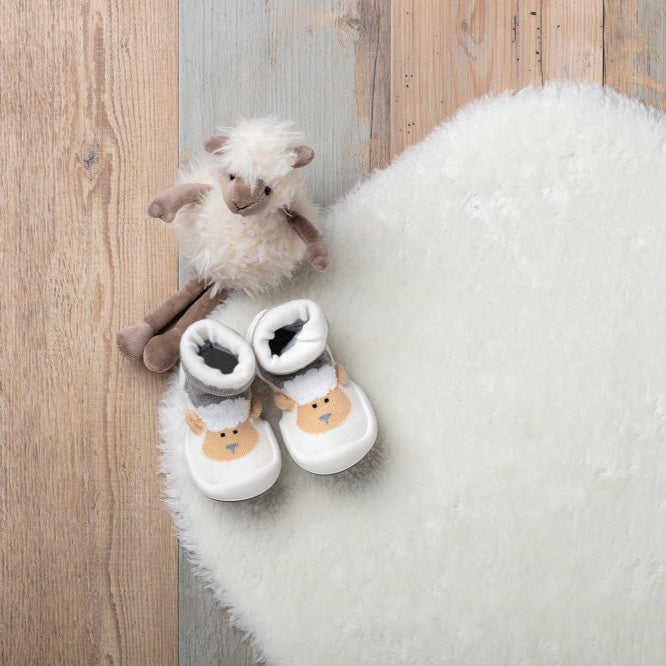 Overhead shot of two sheep-themed baby socks, a stuffed sheep toy, and a white fluffy rug on a wooden floor.