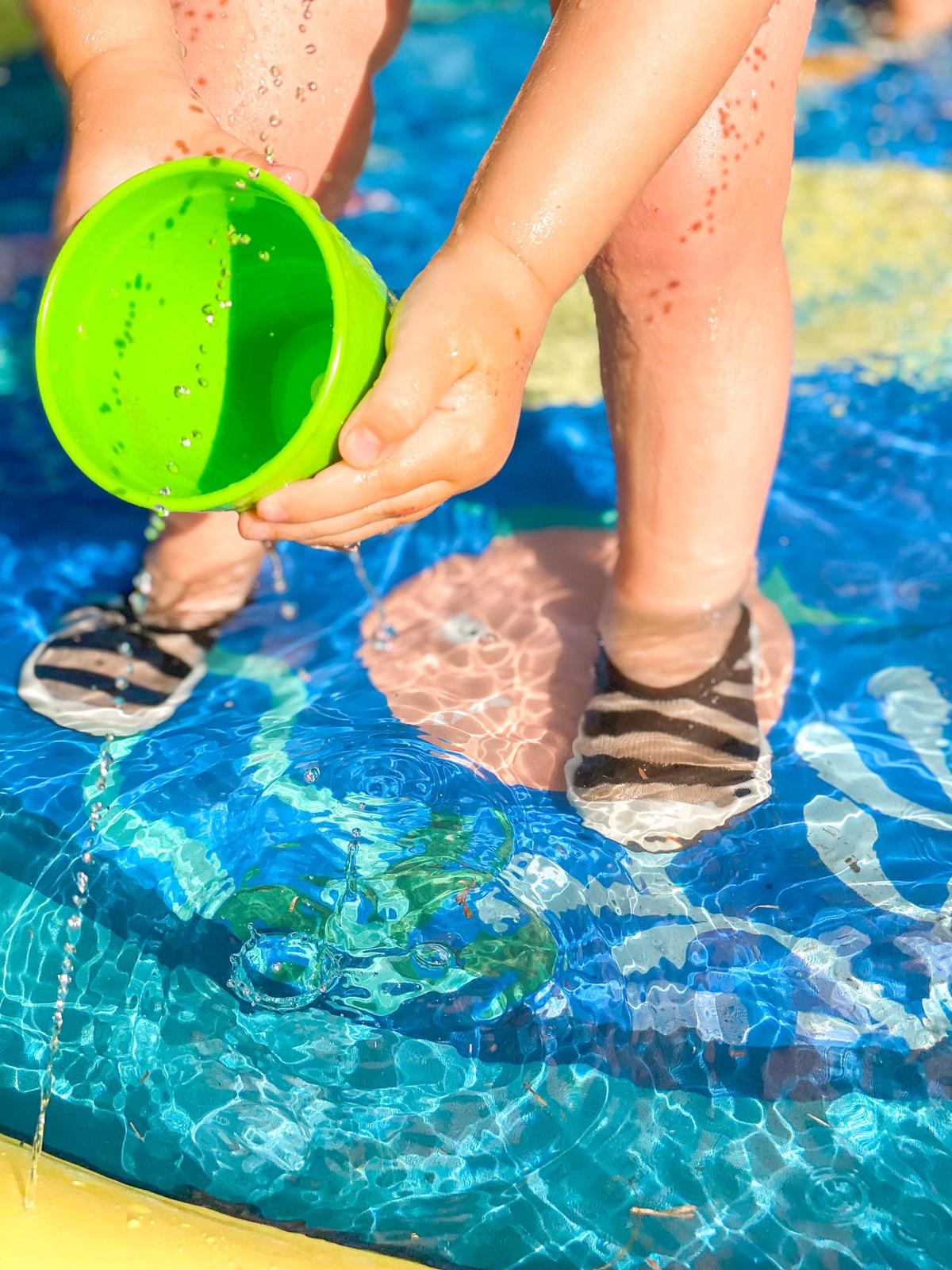 A child stands in a colorful wading pool, pouring water from a green cup. They wear striped swim shoes.