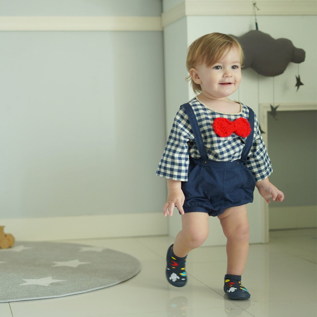Toddler in a blue and white checkered shirt, blue shorts with suspenders, a red bow tie, and patterned shoes, walking in a room with a star-patterned rug.
