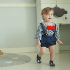 Toddler in a blue and white checkered shirt, blue shorts with suspenders, a red bow tie, and patterned shoes, walking in a room with a star-patterned rug.