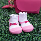 A pair of pink and white baby shoes with bows on a green background with a pink purse.
