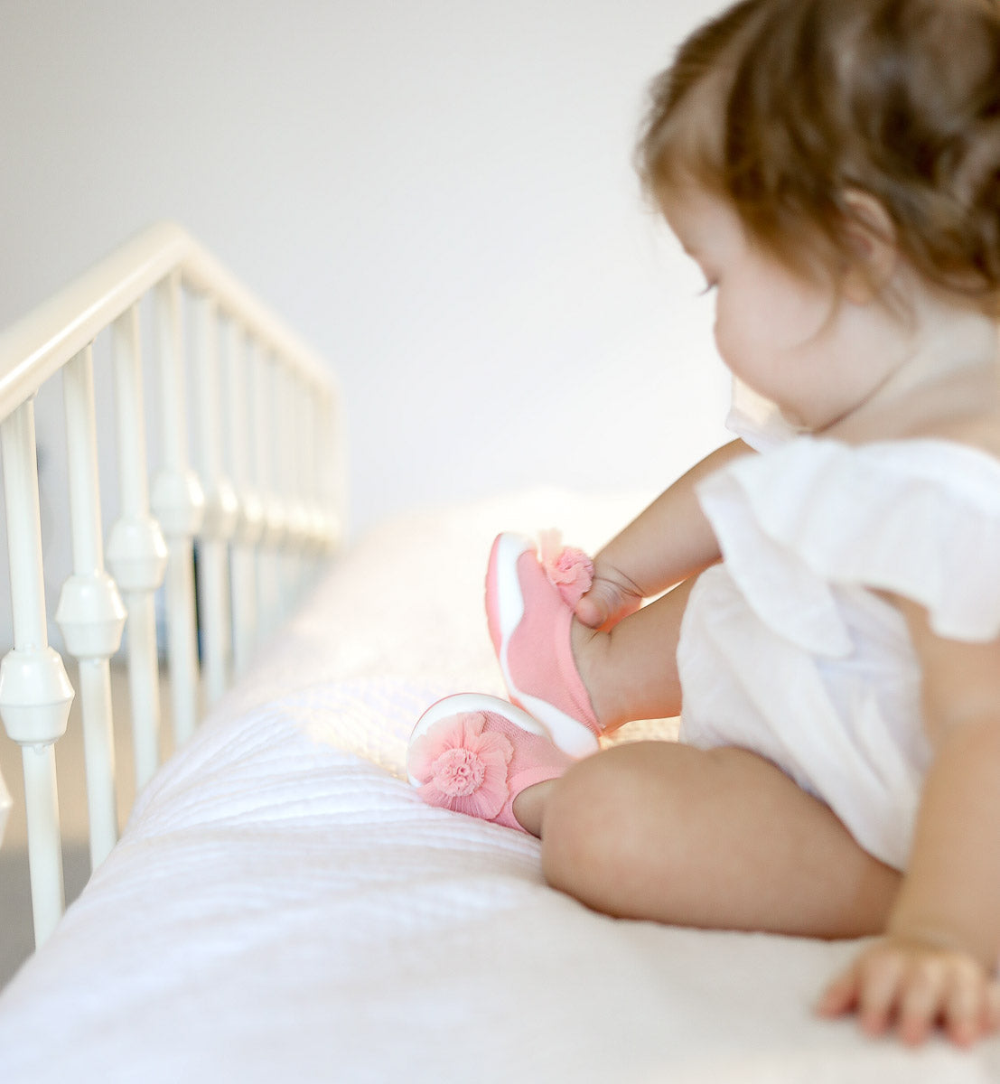 A baby sitting on a white crib wearing pink shoes with flower details and a white dress.