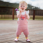 Baby in red striped overalls and pink shoes chewing on a toy outdoors on a wood deck.