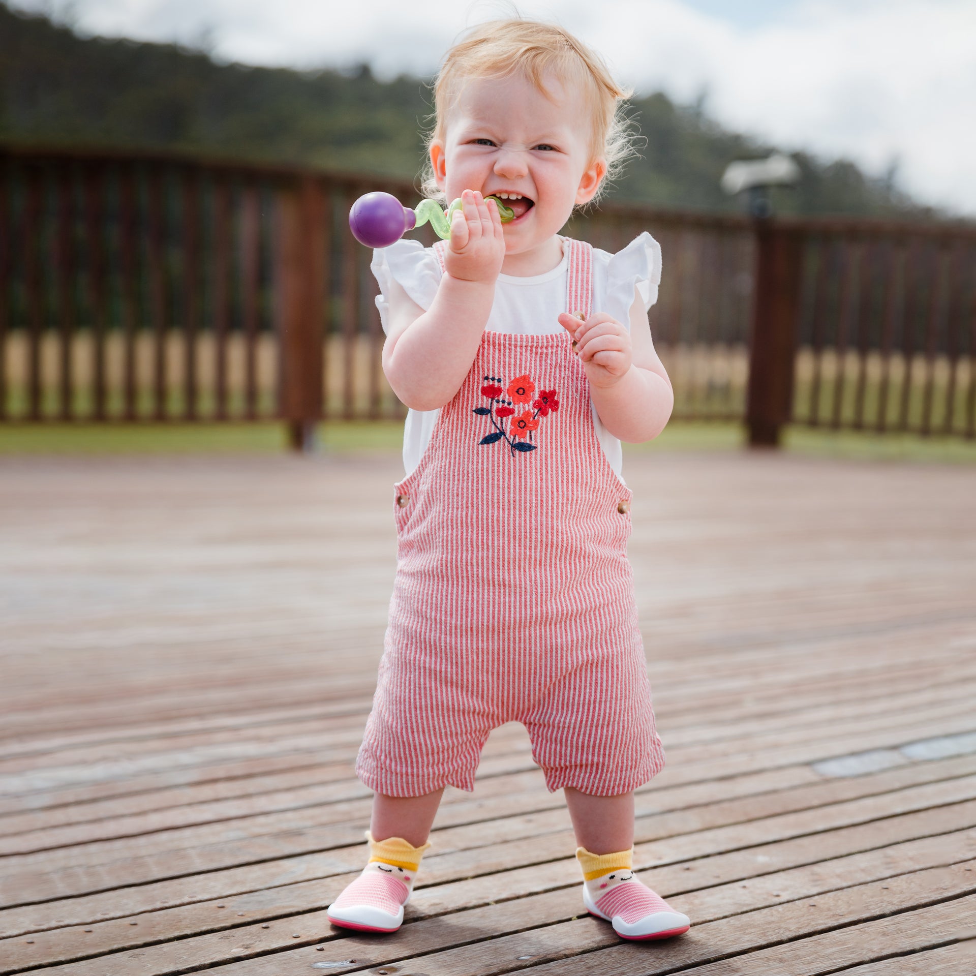 Baby in red striped overalls and pink shoes chewing on a toy outdoors on a wood deck.