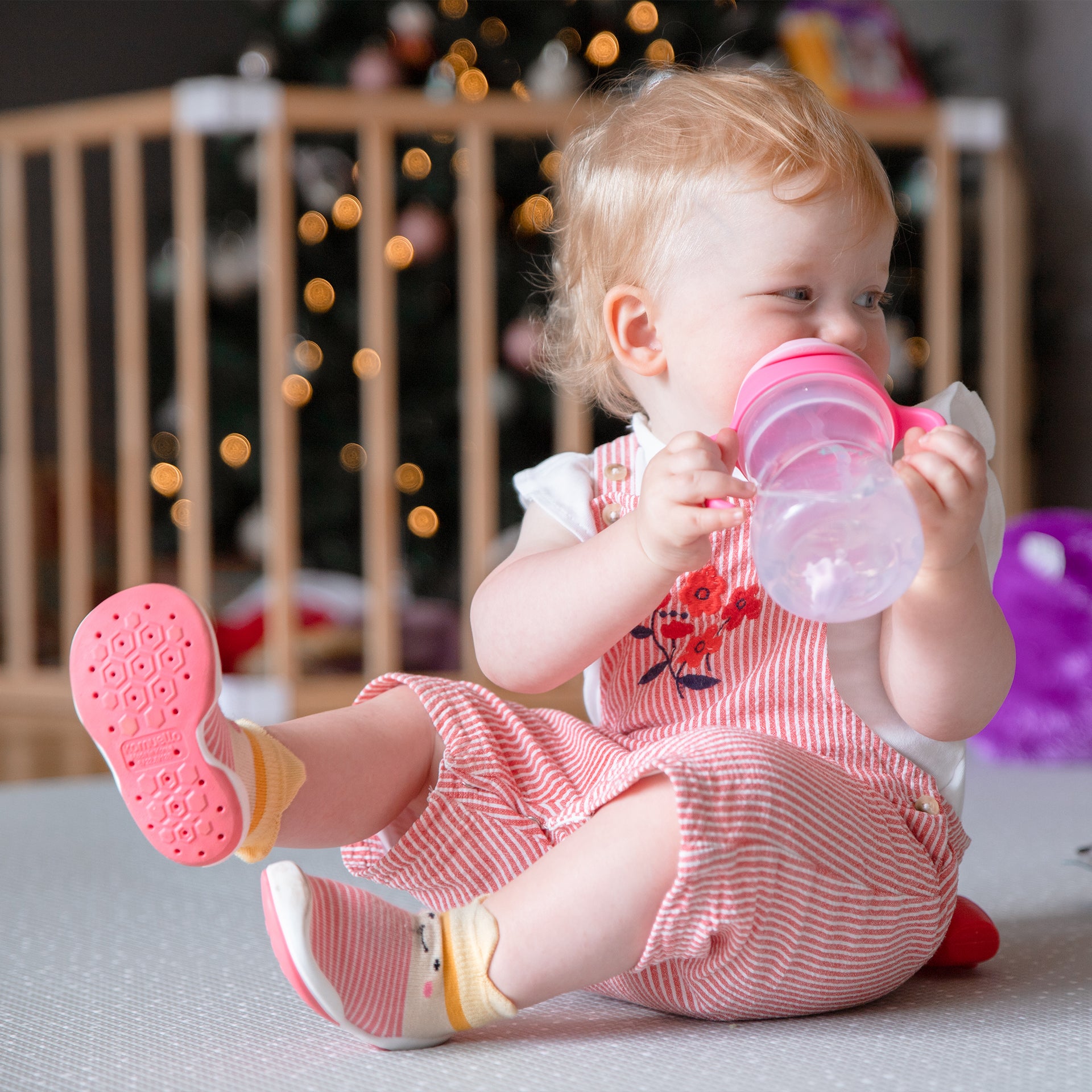 Baby girl in striped overalls and socks drinking from a pink bottle.