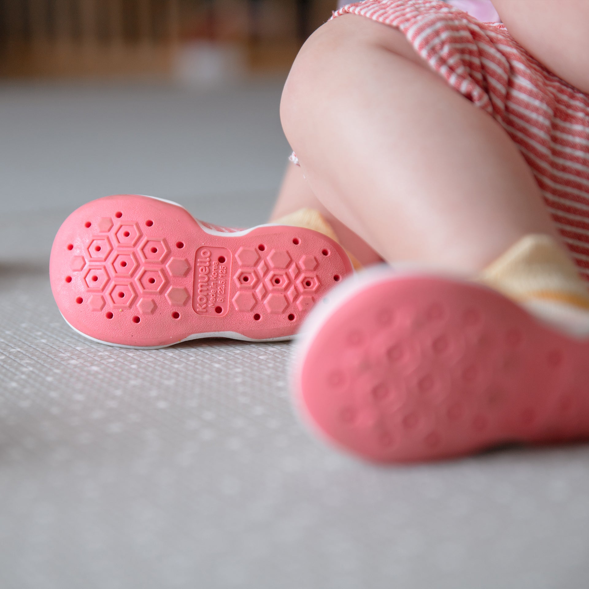 Close-up of baby shoes, pink soles with hexagonal grip pattern, sitting on a gray mat.