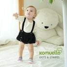 Baby in black romper and polka dot and striped socks, reaching for a bubble, with large white teddy bear behind.