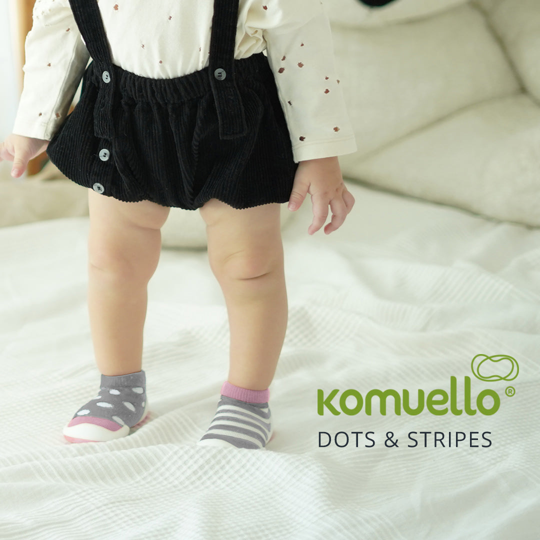 A baby stands on a white blanket wearing mismatched gray and pink socks, black corduroy shorts with suspenders, and a white shirt with brown spots.