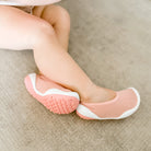 Close-up of a toddler's legs and feet wearing pink and white soft-soled shoes on a textured surface.
