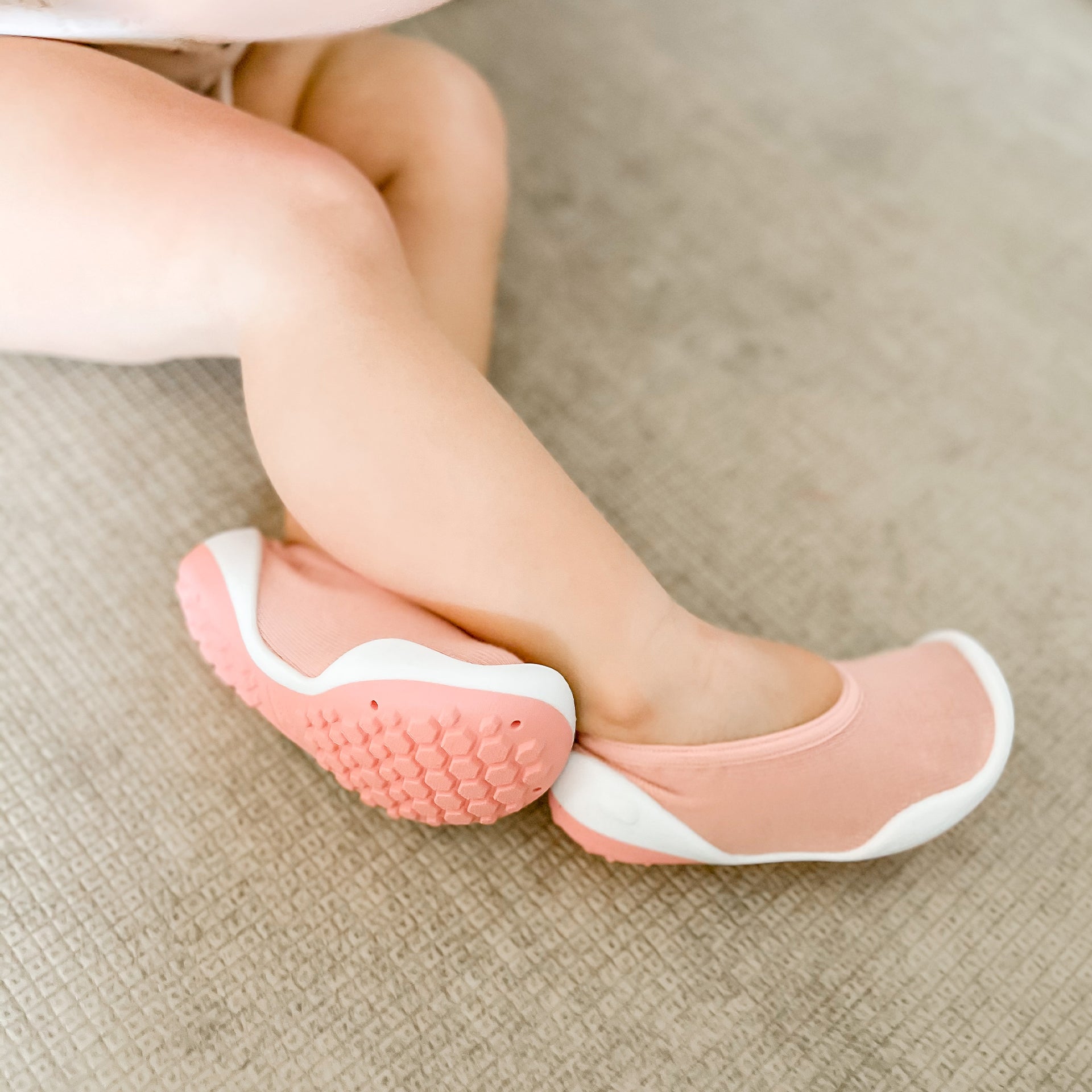 Close-up of a toddler's legs and feet wearing pink and white soft-soled shoes on a textured surface.