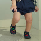 Baby wearing navy blue bloomers, plaid shirt, and dinosaur-patterned socks.