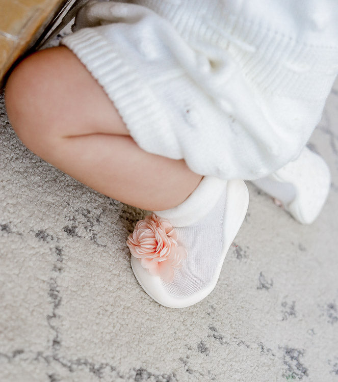 Baby in white cable-knit outfit, wearing white sock-slippers with a peach flower detail.
