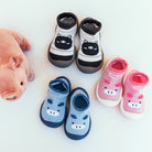 Three pairs of children's slipper socks arranged on a white background, along with a stuffed pig. One pair is white with black stripes and a black cat design, another is pink with a pig design, and the third is blue with a hippo design.