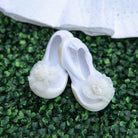Pair of white baby shoes with floral embellishments on a green leafy background.